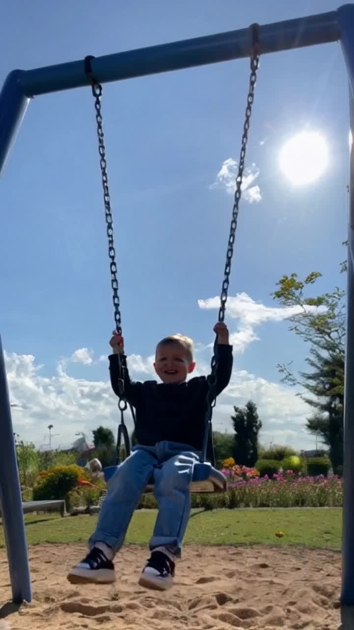 Playful toddler giggling in colorful park swings laughing joyfully while nature blooms around her vibrant sunflowers reaching skyward butterflies fluttering gracefully overhead bright blue sky dotted with fluffy clouds childrens laughter echoing in the background moments of carefree innocence in a serene joyful atmosphere