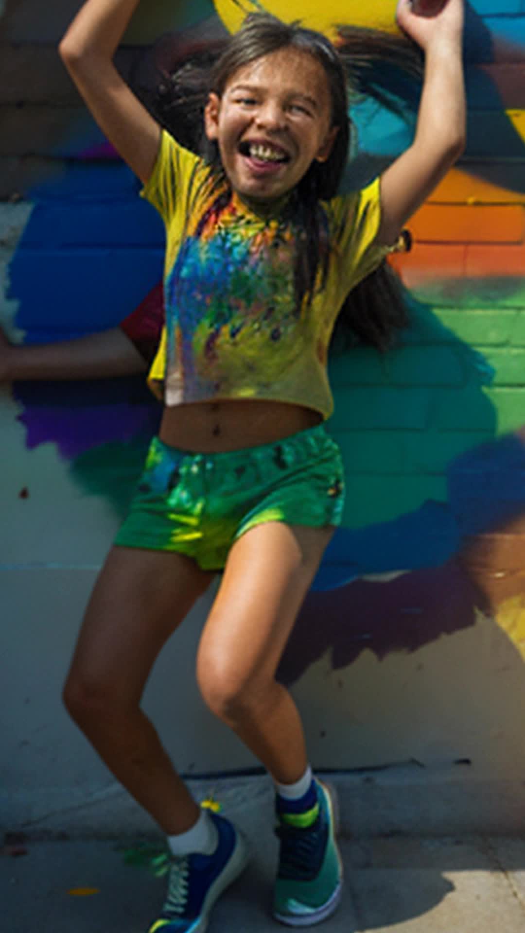 Energetic 10-year-olds splashing vibrant colors, paint-drenched brushes, dull brick wall transforming, quirky local artist guiding, orchestrating dance, drab to vibrant mural, soft shadows, high resolution, wide-angle shot