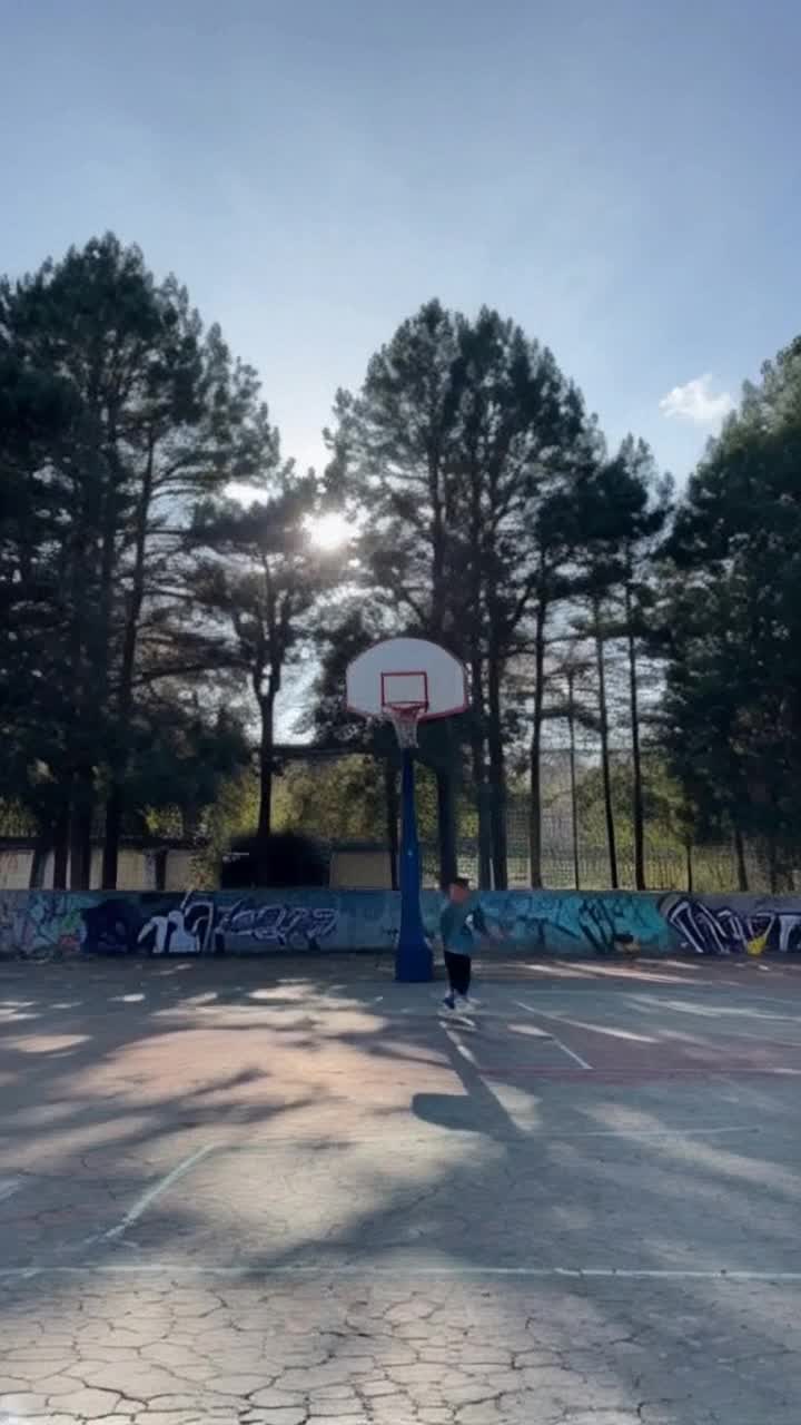Youthful boy dribbling vibrant basketball with fierce determination Bright sunlight casting playful shadows on cracked asphalt court Surrounded by towering trees swaying gently in a warm summer breeze Energetic laughter echoing as friends cheer and shout Nearby graffiti art bursting with color adding an urban flair