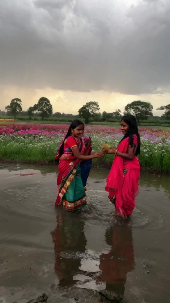 Elegant Indian girls adorned in vibrant saris twirl joyfully beneath raindrops shimmering like diamonds their laughter echoing through the air they leap and dance splashing in puddles forming ripples of joy sparkling water glistens against their colorful attire lush green landscapes surround them with blooming flowers swaying gently in the breeze dark clouds gather above creating a dramatic contrast illuminating the scene with soft, diffused light a hint of rainbow emerges in the distance capturing the essence of blissful celebration amidst natures embrace