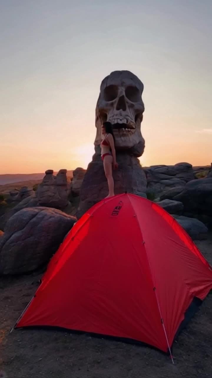 Fancy Gorgeous Instagram Lady Demonstrating her swimsuit, she is shown coming out of a Red tent pitched atop tall boulders near SKULL ROCK in Joshua Tree National Park, the sunset is bold and beautiful, as the scene pans out to show a natural rock formation of a skull, with the red tent on top of it, camping in utopia