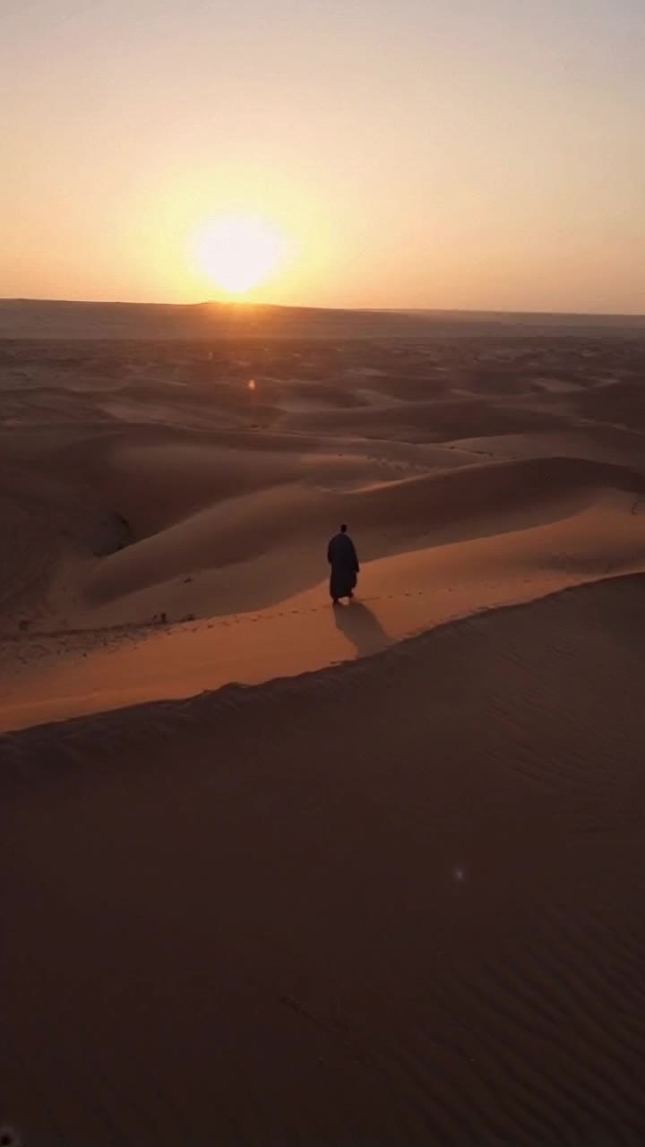 Wide cinematic view of a vast desert at sunset The golden sun hovers low on the horizon, casting long shadows over sand dunes A lone robed man in his 40s walks slowly with heavy steps, his head bowed The wind lifts his robe gently as particles of sand drift in the light