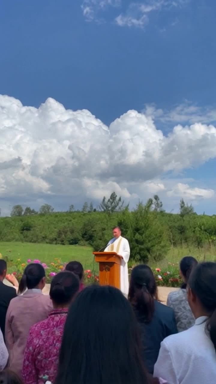 simple white cassock standing at a podium addressing a crowd of eager faces radiating hope and warmth raising hands in prayer under a bright blue sky filled with fluffy white clouds surrounded by lush greenery and vibrant flowers capturing a serene atmosphere of faith and unity