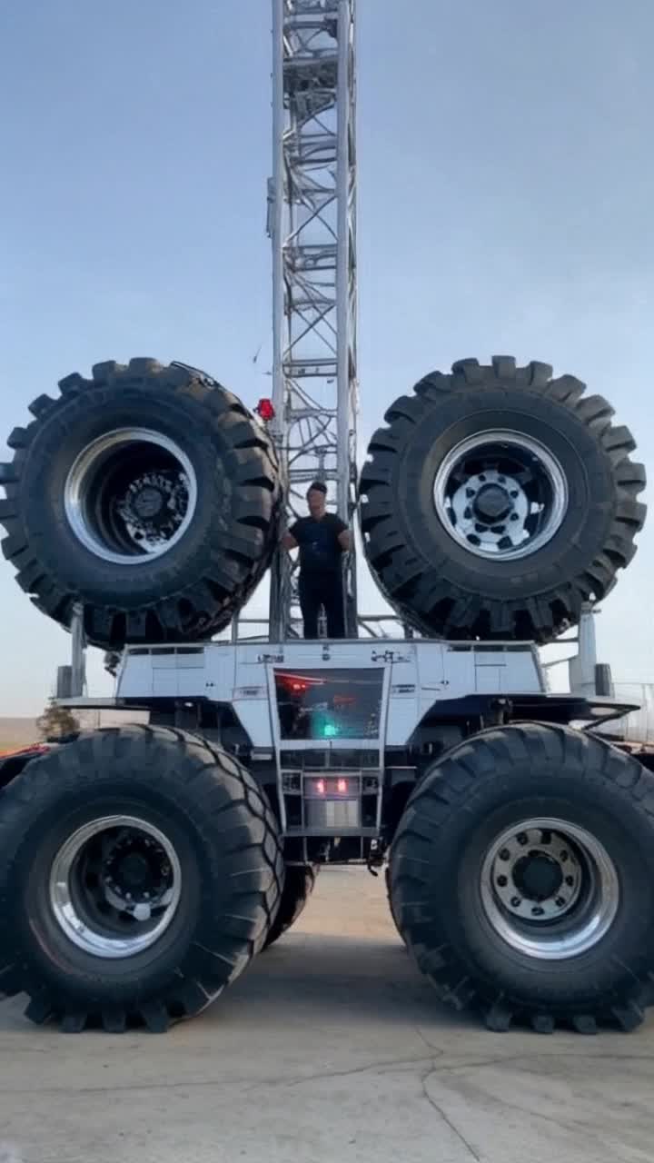 A futuristic monster truck with a raised chrome tower frame The four massive tires are separate, each with its own operator and engine clearly visible through giant glass windows in the stationary central control room of the speeding tires The driver, an impossibly handsome Black man resembling Idris Elba, waves from the window of the trucks main cabin The trucks body houses a show engine for advanced pyrotechnics and laser effects