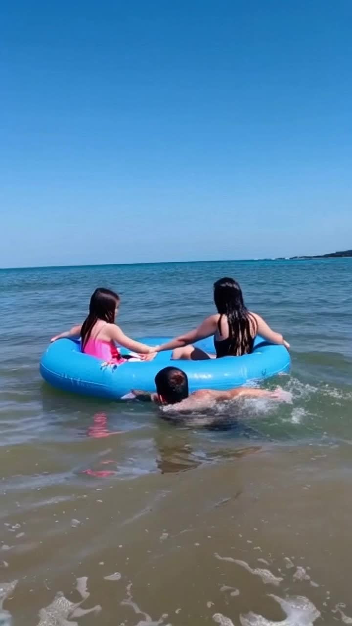 Mother and toddler daughter hold on to opposite sides of an inflatable beach raft , as they relax in the gentle waves they are startled by the sudden appearance of a dark head popping out of the water next to them  The sun is shining and their humorous surprise is comical