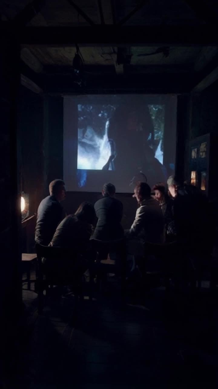 Some intrigued characters huddled closely around an archaic dusty projector curious glances shimmering in flickering light Shadows pulsate embracing their faces Fragmentary images of fully visible faces and figures splayed on screen Gasps resonate as an unseen force beckons them to solve the riddle Hearts race in sync with an eerie soundtrack Outside a sudden thunderous roar disrupts tranquility sending characters into a chaotic frenzy They spring to their feet eyes stoic and wide and resolve ready to confront whatever ominous entity lurks beyond the door Dimly lit chamber swirling shadows heavy whispers of suspense linger in the charged atmosphere