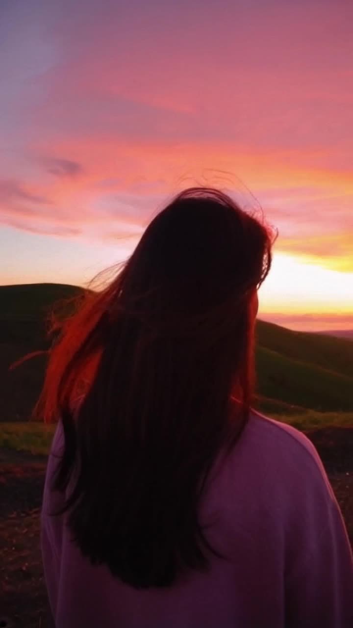 Individual with flowing hair standing confidently against a vibrant sunset Sky painted in hues of orange and pink Surrounding lush green hills gently swaying in the breeze