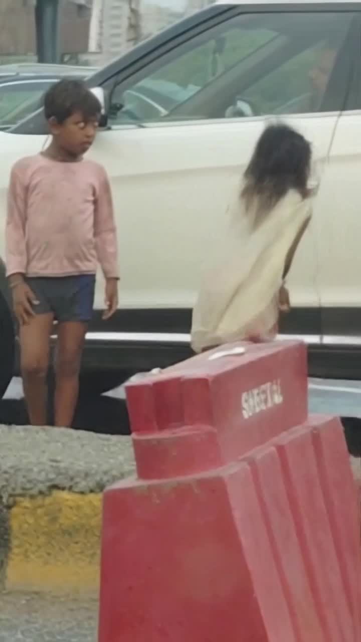 girl wearing  white kurti and red trousers  bouncing joyfully in muddy puddles vibrant splashes of water dancing around her brother wideeyed with surprise droplets soaking his face bustling busy road filled with colorful rickshaws and honking cars gray clouds hanging overhead raining down vivid reflections in puddles creating ripples of laughter and innocence in harsh surroundings