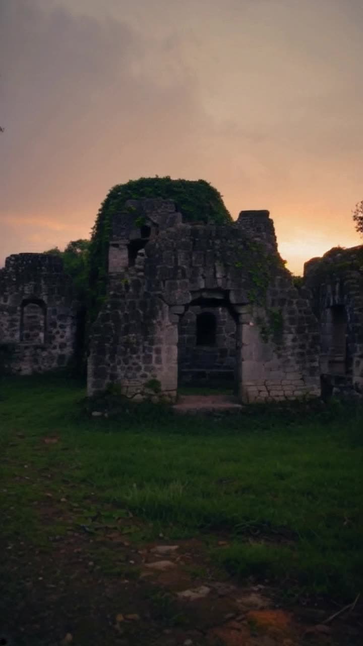 Ancient grey stone structures clustered together exuding charm and character portraying times embrace Eerie silence enveloping the scene as soft winds Lush green grass sprawling around the manor contrasting with weathered stones Overgrown ivy cascades down walls creating a tapestry of natures reclaiming A hazy evening sky painted with oranges and purples sets a wistful backdrop as shadows elongate across the landscape inviting exploration into forgotten stories of history hidden within these quaint relics
