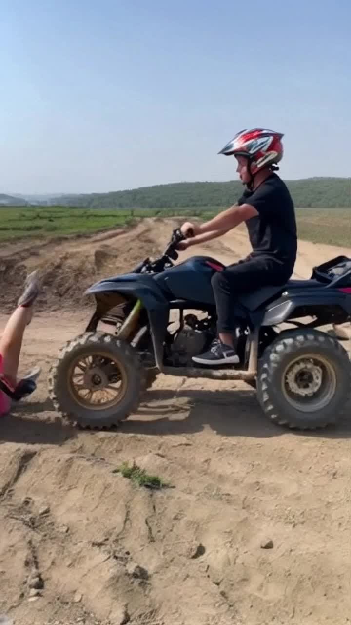 a man wearing a helmet is riding a quad with a woman sitting behind him shes holding onto his waist and shes carryng a handbag trying to show off he starts doing wheelies but he loses control her body scrapes along the ground before she finally falls off the quad