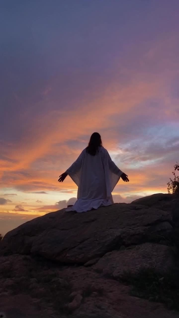 Radiant figure of Jesus Christ with outstretched arms and a serene countenance kneeling on a rocky hillside His eyes closed in deep communion with the divine golden rays of sunlight breaking through dark clouds illuminating his form soft sounds of distant birds chirping and rustling leaves creating a peaceful atmosphere surrounding him vast open skies blending into rich hues of orange purple and blue as dusk approaches