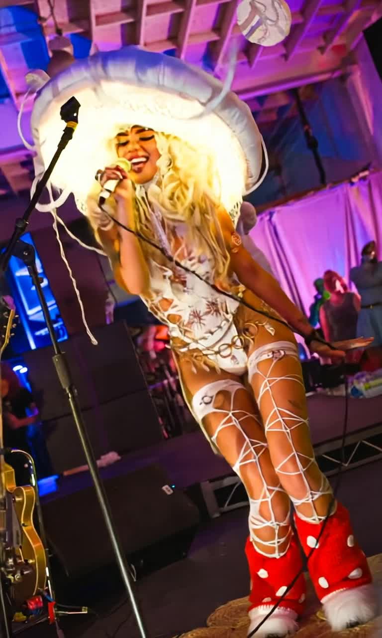 Vibrant woman twirling gracefully under dazzling stage lights Shaking her hips with infectious energy as she sings an uplifting melody Colorful confetti bursts around her creating a festive atmosphere Vibrant visuals of dancers joining in harmony in the background Rich textures of stage props enveloping the performance in an enchanting spectacle Enthralling audience captivated by her electrifying presence as she radiates joy and passion