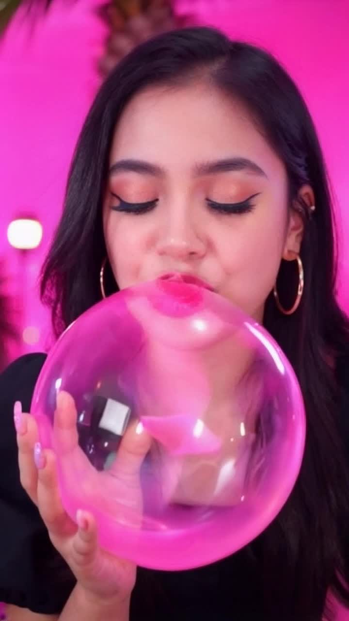 Latina girl in her 20s with long black hair blowing a very big bright pink bubblegum bubble, wearing big thick hoop earrings, black clothes, bright pink background, lights and palm trees in the background