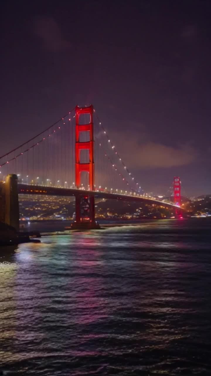 Golden gate bridge illuminated with twinkling city lights shimmering across waters glistening at night glowing reflections