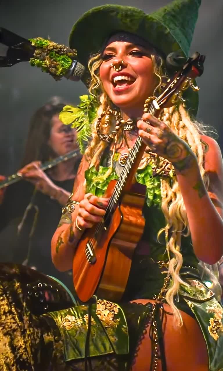 Woman adorned in messy long blond dreadlocks oversized floppy green felt wizard hat costume of vibrant fake leaves shiny green fabric radiates joy strumming a small ukulele with an eartoear grin showcasing her septum piercing on stage enchanted by sparkling fairy lights Soft focus musicians serenade with flutes enveloped in ethereal mist floating icons of PayPal logos Subscribe Now buttons drift around her blending whimsical magic with subtle commercialism An immersive realm where mystical realism dances with vaporwave parody warm glow highlights oversized jewelry cartoonishly bright eyes inviting viewers into this captivating performance