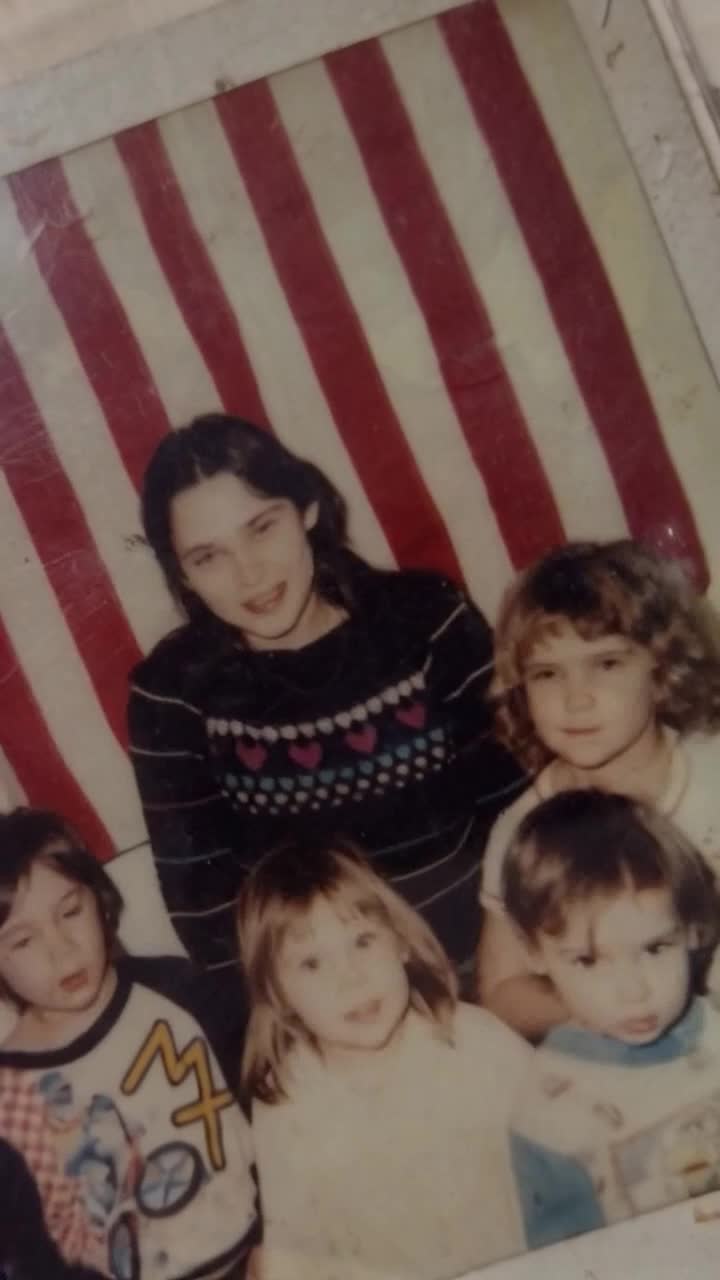Mom is standing in the very back in a dark sweater and there are four children gathered around her all the children jump up with joy and smiles on their face and give Mom a big hug