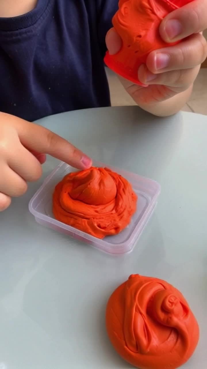 A close up view of child using PlayDoh brand clay mold plastic presses on his little plastic table made for children, bright clay colors as his hands form a red tyrannosaurus and orange triceratops that he pokes at to see if they have dried and are ready to play with