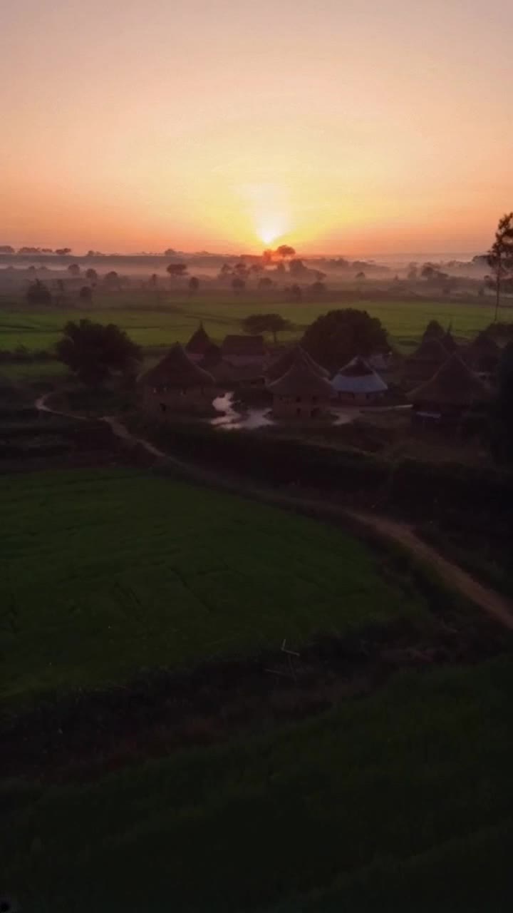 A peaceful rural village at sunrise, with small mud houses, chirping birds, a narrow dirt path, and green fields surrounding the area The sky is tinted with soft orange hues