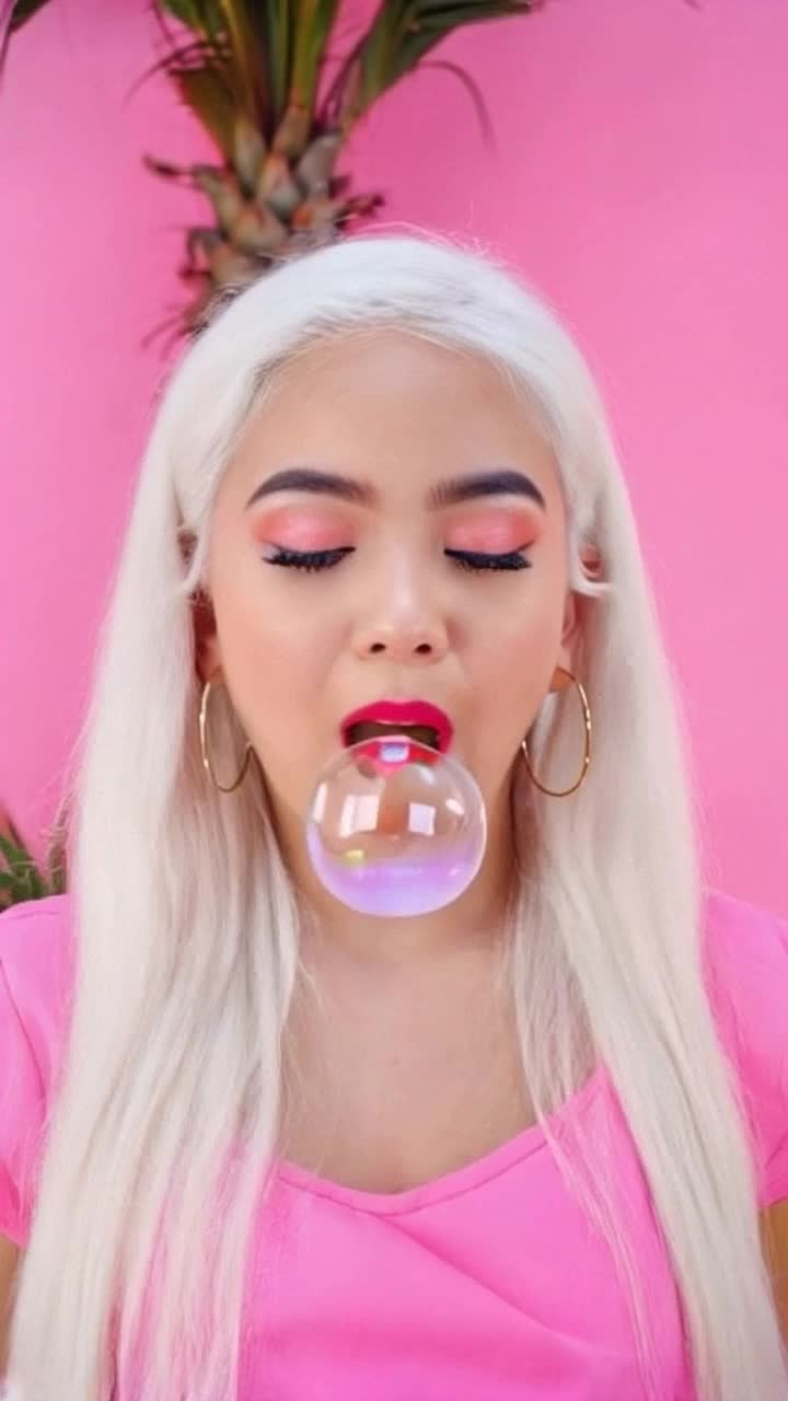 Latina girl in her 20s with long white hair blowing a very big bubblegum bubble, wearing big thick hoop earrings, pink clothes, pink background, palm trees in the background