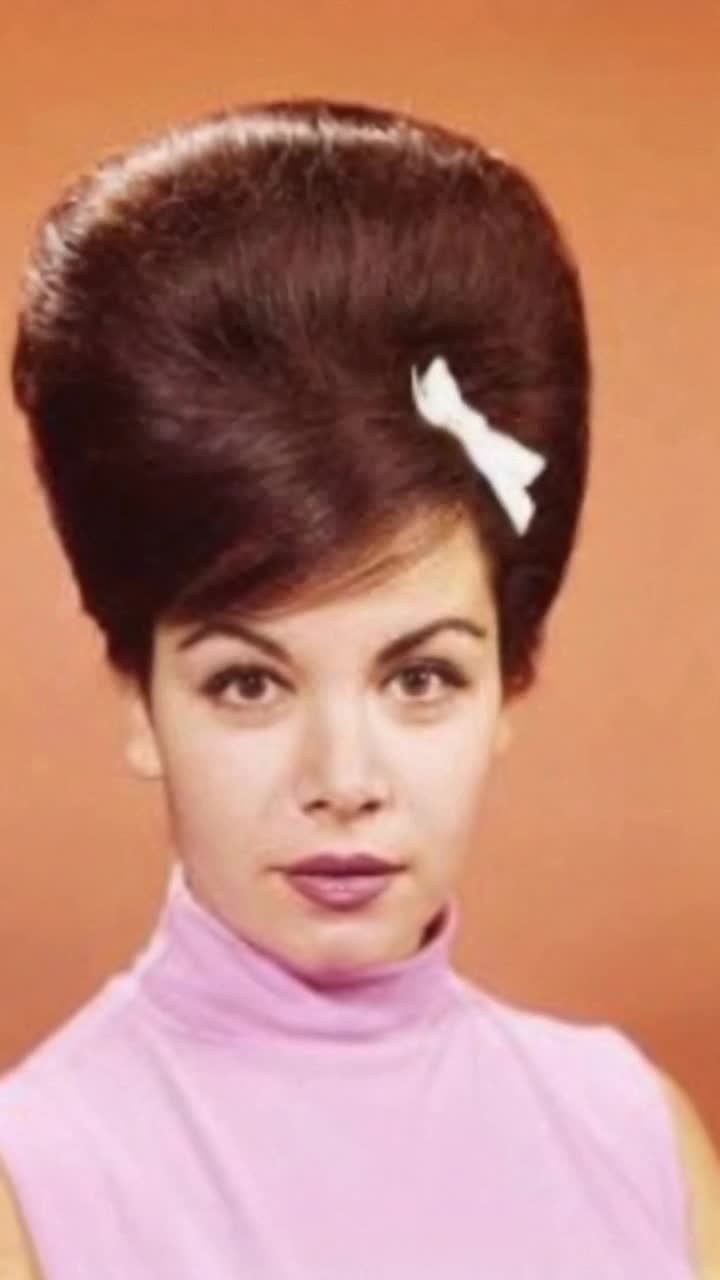 Dressed in a simple 60s outfit of black skirt and white blouse, she gently pats her hair with a fine white gloved hand as  she clasps her purse and turns to look at her reflection in a hall mirror before going out