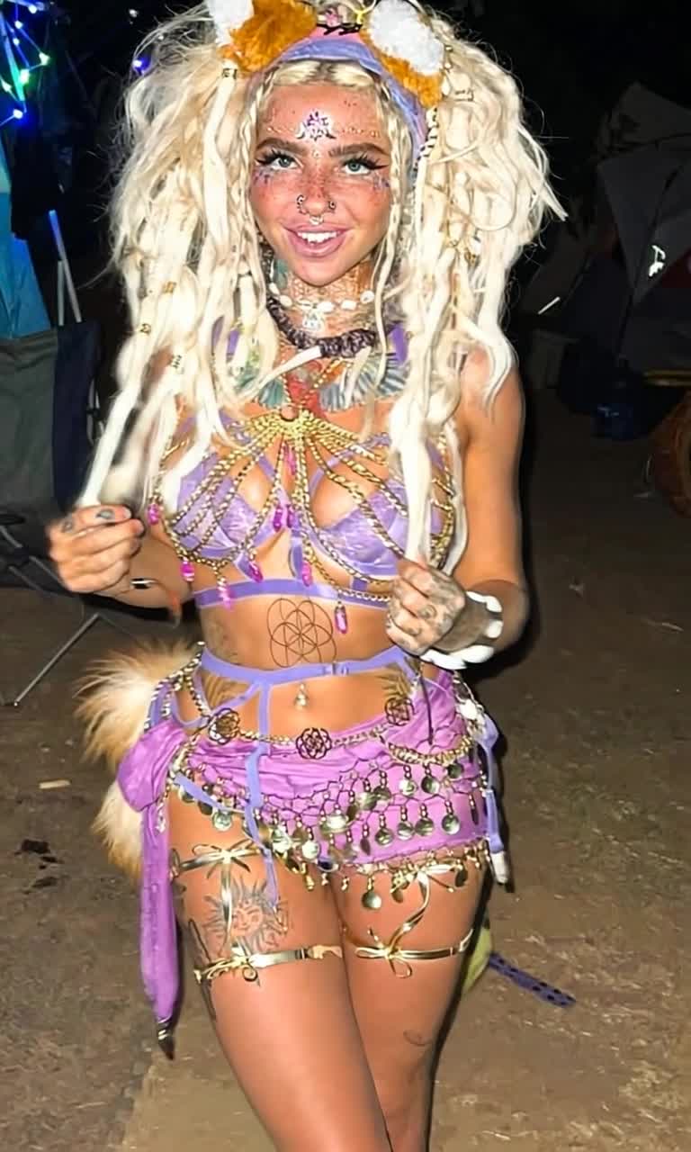 Woman with bleached dreadlocks fox ears glittery face stickers strikes a pose at night twinkling LED fairy lights illuminating her figure purple outfit crafted from ribbons chains coin belts shimmers like a fantasy festival costume Body adorned with random tattoos gold bands reflecting light Forced smile reveals multiple nose piercings Dusty festival campsite unfolds behind her tents folding chairs faded into shadows Tip jar marked Ascension Donations hidden among details dollar signs woven into tattoos Surrealist poprealism style harsh flash lighting creates stark contrast capturing vibrant eccentricity
