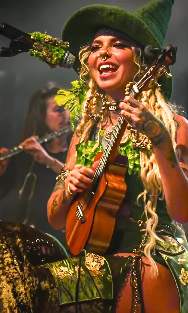 Woman with long messy blond dreadlocks adorned with a floppy green felt wizard hat wearing a costume of glimmering green fabric and vibrant fake leaves strumming a small ukulele with exuberant joy showcasing a wide smile highlighting a septum piercing enchanting audience with playful energy soft focus musicians in the background play flutes surrounded by swirling misty lighting creating a dreamlike atmosphere blending spiritual theater with whimsical charm delicate fairy lights twinkle overhead casting warm glows exaggerated oversized jewelry sparkle as cartoonishly bright eyes shine with delight embracing mystical realism and vaporwave parody